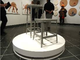 Two gray stools, one larger than the other are on a circular white turntable. Paintings are on the wall in the background.