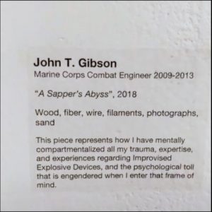 Black printed, sans serif text on a white background reads: John T. Gibson. Marine Corps Combat Engineer 2009-2013. “A Sapper’s Abyss”, 2018. Wood, fiber, wire, filaments, photographs, sand. This piece represents how I have mentally compartmentalized all my trauma, expertise, and experiences regarding Improvised Explosive Devices, and the psychological toll that is engendered when I enter that frame of mind.