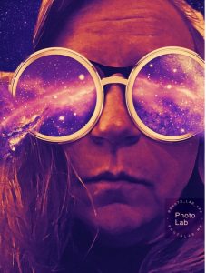 Stylized selfie of the author in a violet purple hue, unsmiling, wearing artistically rendered glasses that have circular lenses with an outer space background. Starry, purple nebulae are exploding out of each lens to the left and right. In the lower right-hand side is a semi-transparent watermark with #PHOTOLAB and photolab.me.