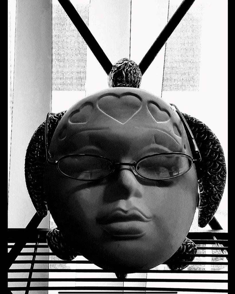 Digital BW Photo, 2019. Depicting The Artist's ambition to connect with The Inner Turtle. A wire shelf supports ceramic mask of contemplative woman's face wearing glasses. Directly behind is a silver sea turtle. Only its head, flipper, and tail are visible as they frame the woman's face.