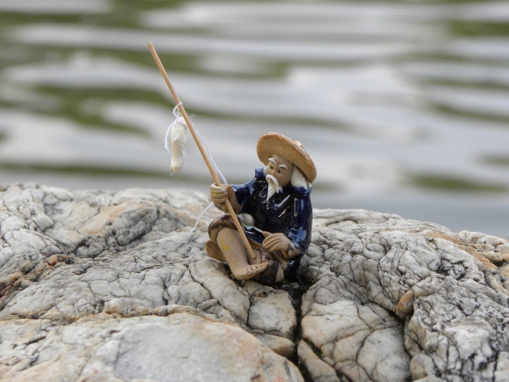 The fisherman figurine with the water in the background. This photograph is divided into two sections. At the center of this picture is a big rock on which a fisherman sits. A tranquil lake is present as part of the background. Dimensions: 1.2 inches × 1.2 inches × 1.2 inches.
