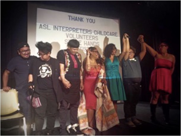 Color photograph of several Crip Your World performers / event participants standing on a dark stage. Behind them is a projected slide that reads, “THANK YOU ASL interpreters, childcare, volunteers,” with the remainder of the listed text unreadable, as the individuals are standing in front of the screen. Three of the people at stage left (right in the image) are holding each other’s hands above their heads.