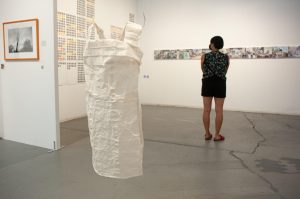 An installation view of the exhibition, Sutured Resilience, showing a corner of the Artworks gallery space. A person stands right of the center to show scale. The foreground image is an ivory paper dress with straps, handmade kobo paper, wire, matte medium, suspended, by Kat Cope. In the background to the left on a perpendicular wall is a black and white photo by Jennifer Cabral of trees and birds. To the right of it on the next wall is, “Birdclock,” 2021, archival pigment print, 80” x 20,” a grid of bird colors categorized by month. The back facing wall is, “MINE_IRA, 2019, pigment print, 11” x 14” x 2”, a series of colored photographs that merges family photos and text, referencing Brazilian mining operations, environmental crisis and, child sexual abuse by Jennifer Cabral