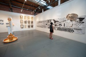 An installation view of the exhibition, Sutured Resilience, showing a corner of the Artworks gallery space. A person stands right of the center to show scale. On the left is a three-tiered sculpture, seven feet tall, Pharma-Go-Round, 2021 by Chanika Svetvilas, with gold mirrored discs and cylinders adorned with various sized melted prescription bottles. Crowning it above is a small chandelier made with prescription bottles dripping with gold chains with one within a brass beaded spiral with hair cascading down from it. Towards the center is a grid of charcoal drawings, “What I have learned (Fill in the Blank),” 2021, each three feet by two feet of text and gaping mouths with various objects inside. To the right, the figure gazes at an 18 foot by four foot charcoal drawing, “KnoWords,” 2021, of text and images on pages of a discrimination case file, Svetvilas vs. The Ohio State University.