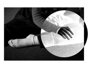 A black and white photograph by Jennifer Cabral. The image shows an arm and leg of a teenage girl sitting on the floor wearing a long-sleeved shirt, black pants and white socks. Her right hand is resting on her leg. Her left leg is bent and her toes are pointed. A large circle is placed over this area of the photograph reversing the image as if it was a film negative. 