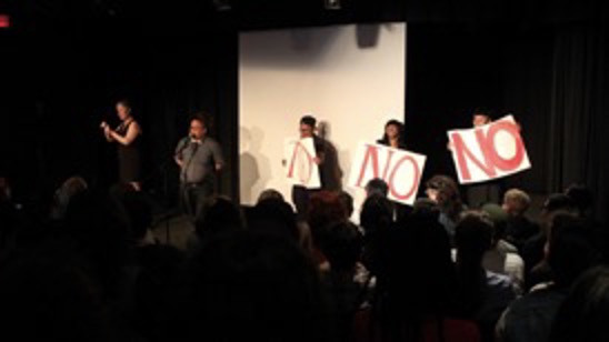 Color photograph of the opening of Crip Your World. A dark stage with five people standing in front of a blank white screen. From left to right are an ASL interpreter, a person at a microphone with their arms behind their back, and three others holding posters with the word NO in large red letters.