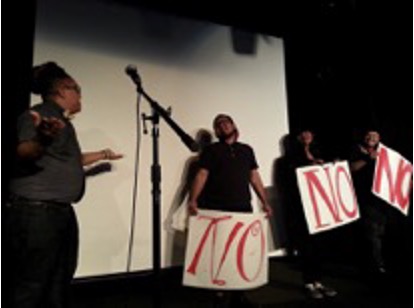 #2 Color photograph of the opening of Crip Your World. A dark stage with four people standing in front of a blank white screen. On the left is a person adjacent to a microphone who is gesturing with their arms, facing three others to the right on stage who are holding posters with the word NO in large red letters.