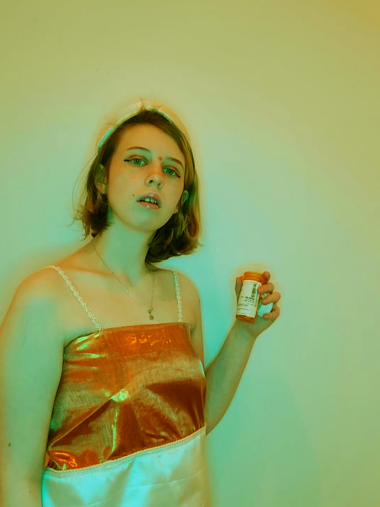 A series of three color photographs depicting the artist wearing an orange and white dress resembling a pill bottle. They have on a matching white hat that is shaped like the cap of a pill bottle. The first photo they are facing to the left of the screen and have their body pulled inward, not wanting to acknowledge being looked at and trying to make themself smaller. In the second, they are staring into the camera holding up a small plastic pill bottle in their hand. The final shows them looking up and to the right while leaning forward with an annoyed expression. Artist Sidne K. Gard took the photographs, as well as fully sewed and designed the outfit.