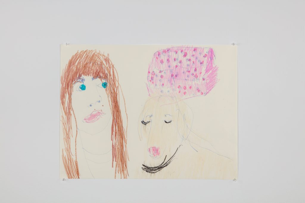 “Self portrait with London and couch”. The artist, Emilie is on the left. She has long brown hair, blue eyes, pink lips, and is wearing a white t-shirt. London looks at an angle down to the right side of the drawing, and the couch floats above her head.