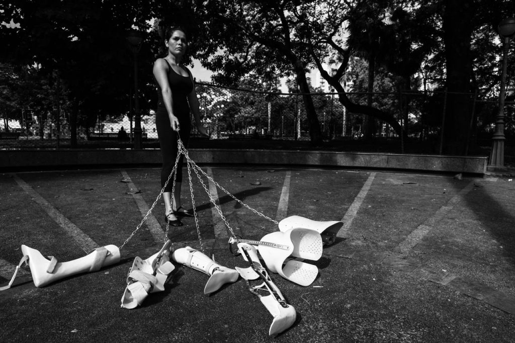 Black and white image of my performance Poética Prótetica (Prosthetic Poetics). I am in one of the main squares in the city of Salvador, Brazil. I hold with my right hand a variety of body limb orthotics attached by steel chains. They seem like dogs taken for a walk. I am wearing black dance clothes and ballet slippers. And my face has a strong expression looking forward. In the background, many trees and the protective fence around the square.