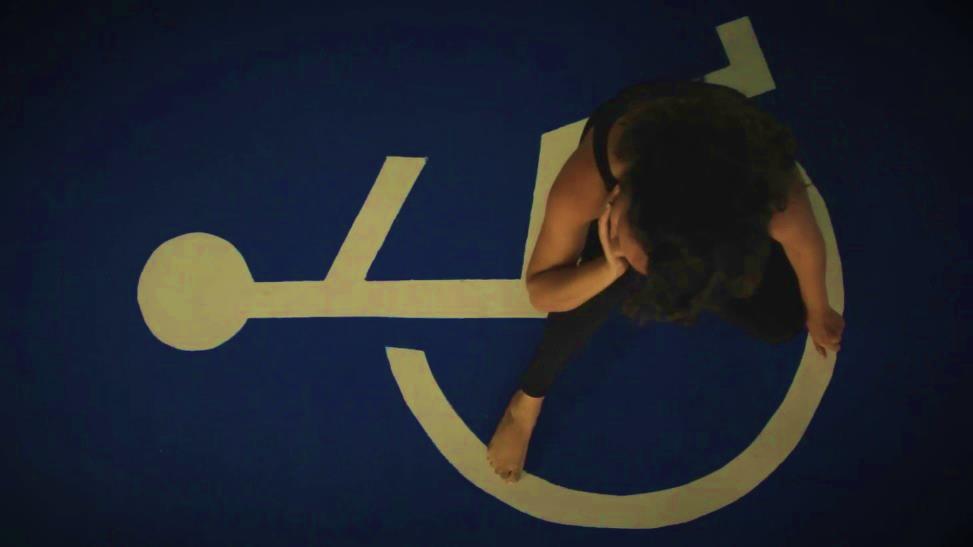 A zenithal view of a film Carolina by the director Lilih Curi (Brazil). The image shows a zenithal or aerial view of the short film Carolina, by the director Lilih Curi (Brazil). I am sitting under a huge parking space for people with disabilities and I dance specifically on the international symbol for accessibility. I am in a thinking position, with my right hand supporting my chin and facing the wheelchair user's drawing on the floor.