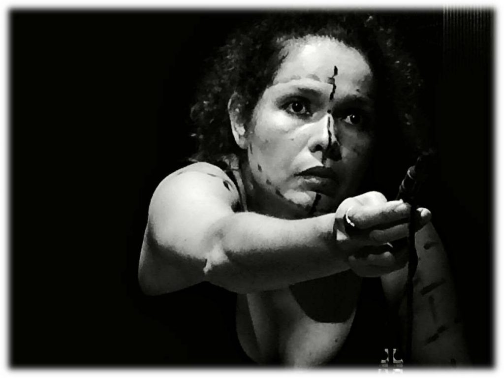 A black and white photo taken in a theatre in Brazil. I am on stage, sitting under a central spotlight. My face is streaked with small black lines that divide me. My eyes and lips are painted up in black. I'm staring at the audience in supplication, in order to offer the spectators a pen to finish streaks on my body.