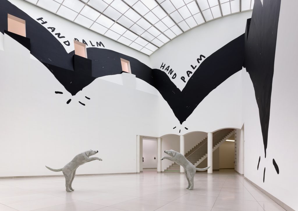 “Crip Time, Dancing with London” A bird’s eye view of a gallery space, with a sculptural installation of “Dancing with London”, with a black and white mural painted on the wall, “Hand Palm” by Christine Sun Kim.