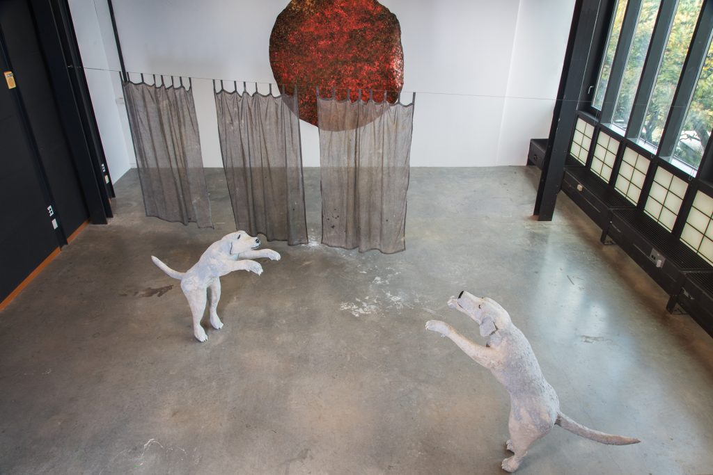 “Looking at the Sun and Dancing with London.” An installation shot from above of the artist’s studio with white walls, big windows, and a gray concrete floor. The installation is of two larger than life sculptures of a female Labrador Retriever standing upright on their hind legs, with their paws outstretched towards each other, as though they are leaping up into the air to dance with each other. There is about 5 feet of distance between them. To the left, are three semi-transparent curtains hanging from the ceiling, separating the sculptures from a large, circular orange tapestry, that also hangs mid air from the ceiling. It is displayed to look as though the sun is partially hidden behind the curtains. The mood in this space is happy, joyful, and bright.