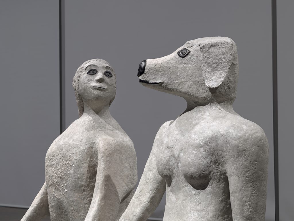 “True Love will Find You in the End.“ Two sculptures depicting hybrid human-animal figures stand side by side in a gallery space. They are seen close up, from the shoulders up, and from a slight profile view. On the right, is a naked woman with a dog’s head; the figure on the left is an anthropomorphized dog with the head of a woman. Both of their features are very stylized, with big, almond shaped eyes, and well defined eyelashes painted black.
