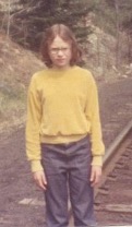 Blurry photograph of the author at 10 years old with shoulder length brown hair, squinting at the camera, wearing glasses, a gold long-sleeve velour shirt and bluejeans. I am standing on railroad tracks and behind me are brown weeds and a green pine tree.