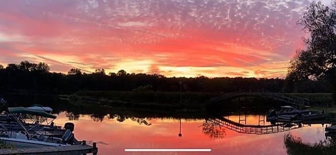 A sunset reflected off an inlet off near Black Lake in Central New York. There are wisps of clouds across the top and shades of purple, pink, and yellow are in the sky. Miscellaneous motor boats are on each side and a walkway bridge is reflected in the water.