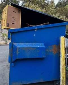 A Dumpster painted bright blue sits next to a yellow concrete pillar. Both show signs of corrosion, red rust on the Dumpster and peeling paint on the pillar. The Dumpster’s lid is held open by a cardboard box. The yellow trim on the box, which is at the upper left, echoes the yellow pillar in the lower right of the frame. The effect is balanced and restful.