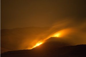 A hilly landscape at night time with hazy smoke and wildfire flames being blown by wind. Image by Kevin Ellis from Pixabay. Free for commercial use. No attribution required.