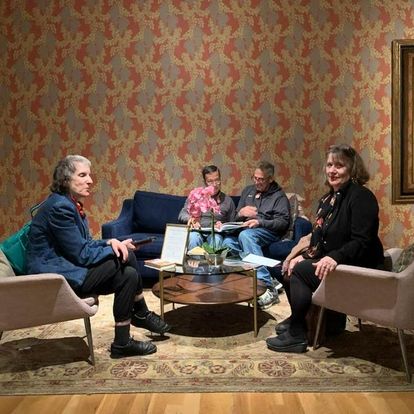 Kathi Wolfe, on the left, and Sheila Black, on the right, sitting in a lounge at the Phillips Gallery. Between them is a flowering plant with pink blossoms on a round coffee table. In the background, two men are sitting on a dark blue couch.