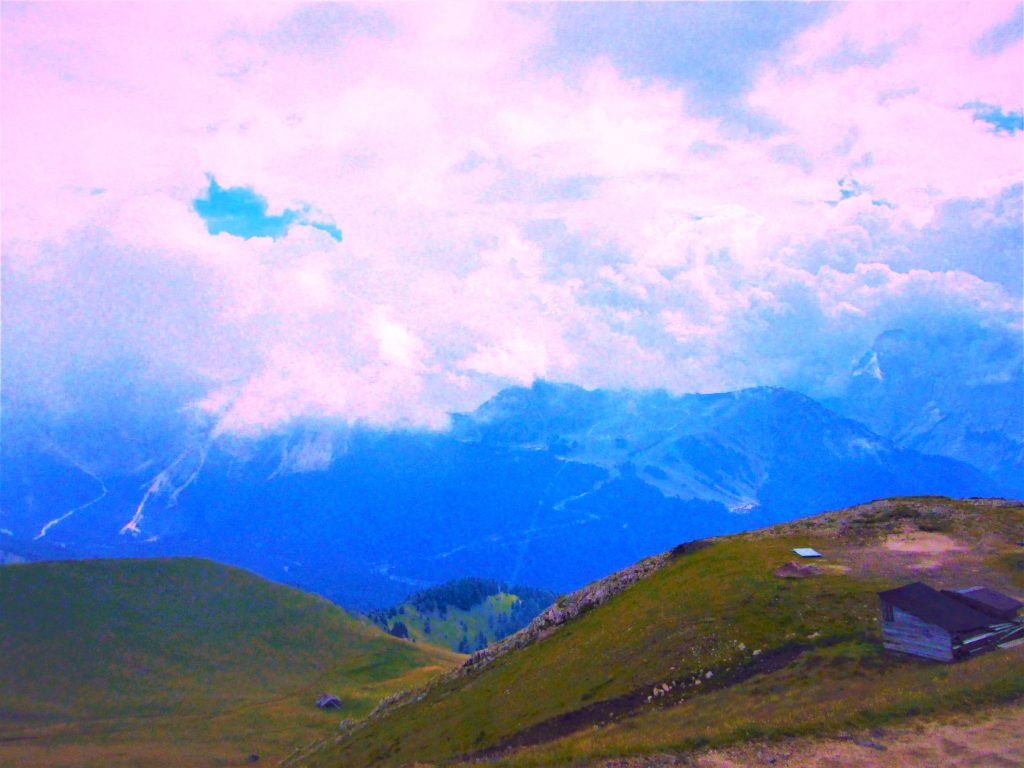 Color photograph taken with a point and shoot camera, edited with Microsoft Word. In the lower right corner, a solitary hut on a green hilltop faces a wide, expansive valley in the Dolomite Mountains. The mountain peaks in the background are shaded blue and there are violet-tinged clouds in the sky overhead.