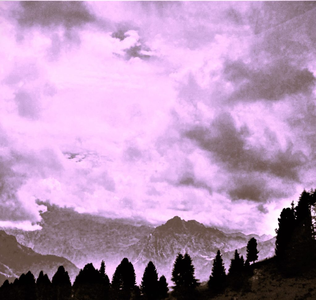Color photograph taken with a point and shoot camera, edited with Microsoft Word. Sunrise in Dolomite Mountains. Several dark trees are outlined in the foreground, with mountainous terrain behind them in the distance. The sun is barely peeking through on the right.. The sky is full of clouds. The entire scene is lilac-tinged. 