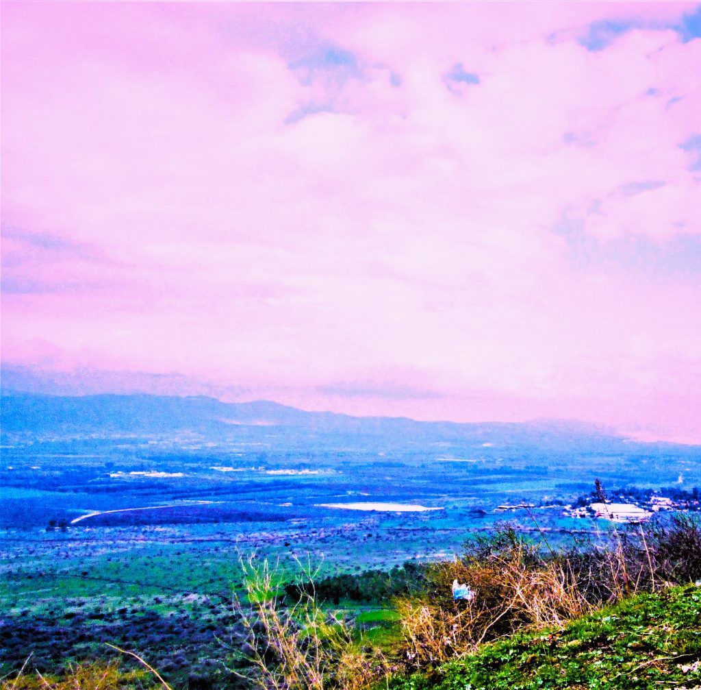 Color photograph taken with a point and shoot camera, edited with Microsoft Word. Flat fields visible from mountains in shades of blue. On the right, a small village is shown. Several ponds appear sporadically. The sky is full of flat, lilac-tinged clouds. In the foreground, greenery and bushes appear.