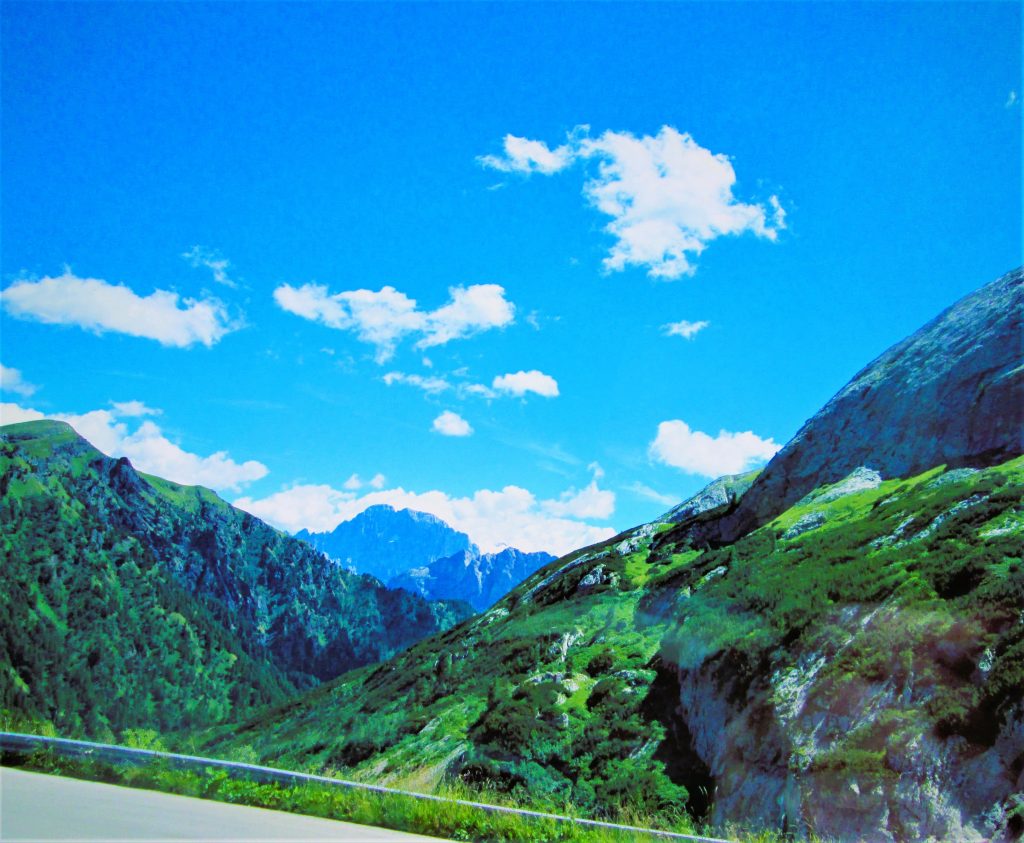 Color photograph taken with a point and shoot camera, edited with Microsoft Word. Mountains covered in green in the Dolomite Mountains as viewed from a roadway. There are peaks in the distance in shades of blue. Variously shaped and spaced clouds appear in the blue sky overhead.