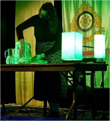 a vibrant green, similar to the color of lime jello, lights up a room and the image. lauren samblanet, a white gender fluid person with long brown hair, wears glasses, a mask, a dark sweater, and a flower skirt. they are standing behind the table on the stage with green lights on it, gently dropping stones into the large glass vase.