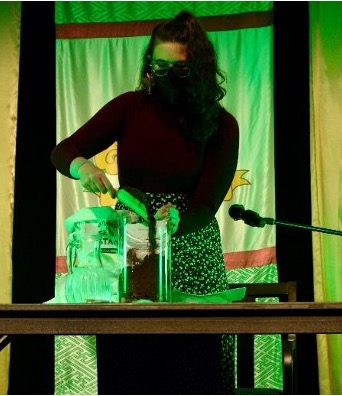 a lime-green tinted photo of lauren standing behind the table with the green lights. she is holding a shovel and shoveling dirt on top of their book in the glass vase.