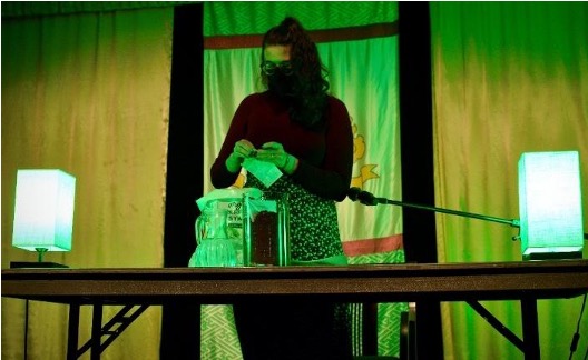 another lime-green tinted photo of lauren standing behind the table with the green lights. they are opening a package of chamomile seeds, so that they can scatter them into the glass vase that has her book and dirt in it.